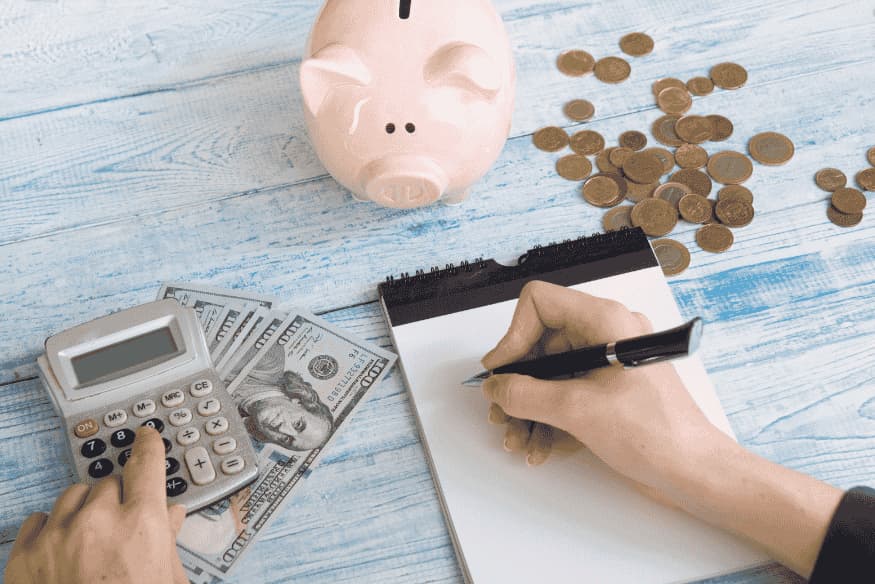 Person calculating expenses and writing a savings plan next to a piggy bank and scattered coins.