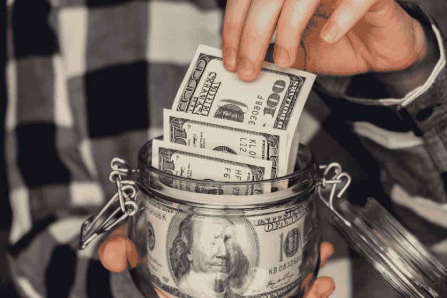 Person putting dollar bills into a glass jar, representing effective money-saving strategies.